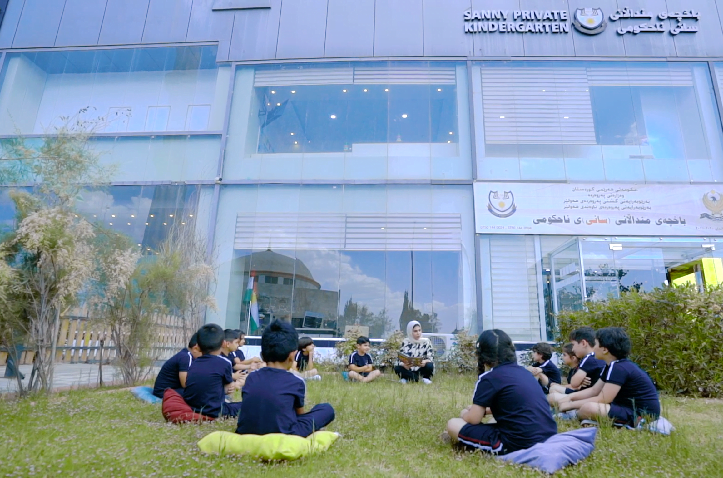a group of students sitting on the grass in front of a sanny kindergarten building.